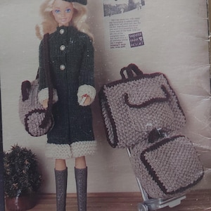 May include: A doll wearing a green knitted coat with white trim, a brown hat, and brown boots. The doll is holding a brown knitted bag and is standing next to a set of brown knitted luggage with a silver handle.