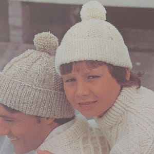 May include: Two people wearing white knitted hats with pom-poms. The person in the front is wearing a white knitted sweater. The person in the back is wearing a white knitted sweater with a cable knit pattern.