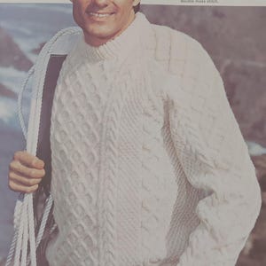 May include: A white cable knit sweater with a textured pattern. The sweater is worn by a person with brown hair and a beard. The text on the image reads "Make Him A Winner", "Materials", "Sirdar Sportswool", "Sirdar Sherpa", "Sirdar Bainin", "1 Pair No. 8 and No. 10 Knitting Needles, 1 Cable Needle", "Tension", "The tension for this design is 10 stitches to 2 inches (5 cms) over double moss stitch."