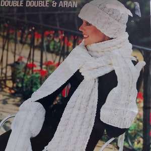 May include: White knitted hat, scarf, and mittens set. The hat has a lacy design and the scarf and mittens have a cable knit pattern. The set is shown on a model sitting on a chair.