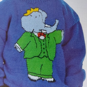 Könnte beinhalten: Ein blauer Strickpullover mit einem Comic-Elefanten, der einen grünen Anzug und eine rote Fliege trägt. Der Elefant trägt eine gelbe Krone auf dem Kopf.