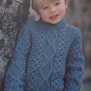 Peut inclure: Un jeune garçon portant un pull bleu en tricot avec un motif en câble. Le pull a un col rond et des manches longues.