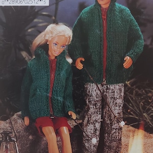 Könnte beinhalten: Zwei grüne Strickjacken mit roten Reißverschlüssen, eine mit Kapuze und die andere mit Kragen. Die Jacken werden von zwei Puppen getragen, eine sitzt und die andere steht. Die Puppen halten Stöcke mit Marshmallows darauf, und im Hintergrund ist ein Feuer zu sehen.