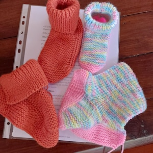 Könnte beinhalten: Zwei Paar gestrickte Babyschuhe. Ein Paar ist orange und das andere ist pink mit blauen, grünen und gelben Streifen.