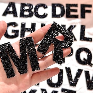 May include: Close-up of the letters 'M' and 'R' covered in black rhinestones. The letters are held in a hand, with other rhinestone-covered letters in the background. The letters are for crafts and DIY projects.