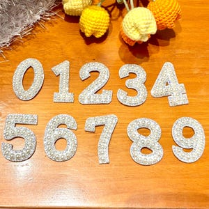 Könnte beinhalten: Eine Reihe dekorativer Zahlen von Null bis Neun. Jede Zahl ist silberfarben, mit einem Rand aus kleinen, klaren Strasssteinen und gefüllt mit Kunstperlen. Die Zahlen sind auf einer Holzoberfläche angeordnet.