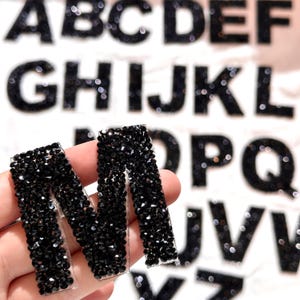 Könnte beinhalten: Nahaufnahme des Buchstabens "M", bedeckt mit schwarzen Strasssteinen, gehalten vor einem verschwommenen Hintergrund anderer mit Strasssteinen bedeckter Buchstaben. Der Buchstabe befindet sich auf einer transparenten Rückseite.