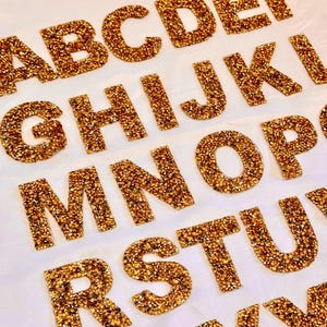 Könnte beinhalten: Nahaufnahme von goldenen Glitzer-Alphabetbuchstaben. Die Buchstaben A, B, C, D, E, G, H, I, J, K, M, N, O, R, S und T sind sichtbar. Die Buchstaben sind auf einer weißen Oberfläche angeordnet. Die Buchstaben sind mit goldenem Glitzer bedeckt.
