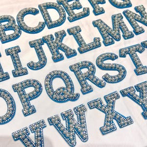 May include: A selection of alphabet letters, each featuring a blue backing and embellished with faux pearls and sparkling rhinestones. The letters are displayed on a white surface, creating a decorative and visually appealing arrangement. Perfect for craft projects or custom gifts.