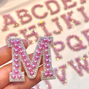 Könnte beinhalten: Nahaufnahme eines rosafarbenen Buchstaben "M" Aufnähers, verziert mit rosa Perlen und klaren Strasssteinen. Der Buchstabe wird in einer Hand gehalten, mit anderen rosa, glitzernden Buchstaben im Hintergrund. Der Aufnäher hat einen weißen Rand.