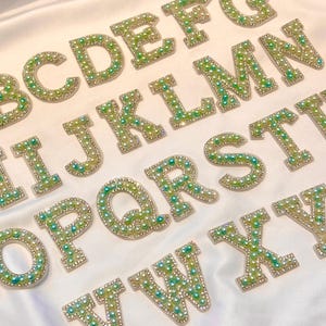 Könnte beinhalten: Nahaufnahme von Buchstaben des Alphabets, die jeweils mit grünen und weißen Perlen und Strasssteinen verziert sind. Die Buchstaben sind auf einer weißen, seidigen Oberfläche angeordnet. Die Buchstaben sind in einer Serifenschrift, und die Gesamtästhetik ist dekorativ.
