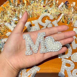 May include: Close-up of the letter "M" and a heart-shaped embellishment, both adorned with faux pearls and rhinestones. The letter and heart are held in a hand, with other pearl-studded letters visible in the background.