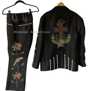 Puede incluir: Traje negro de estilo western con intrincados bordados. La chaqueta presenta un dise&ntilde;o de cruz y cactus en la espalda, mientras que los pantalones tienen motivos florales y de pistola. El texto "Bespoke Addiction" es visible.