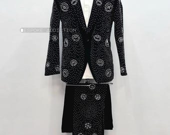 May include: A black suit with white rose-shaped embellishments. The suit includes a blazer and matching pants, displayed on a mannequin. A white collared shirt is visible beneath the blazer. The text "BESPOKE ADDICTION" is visible on the left side.