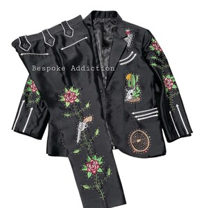 Puede incluir: Traje negro con bordados florales, cactus y pistola. La chaqueta y el pantal&oacute;n presentan detalles blancos y el texto "Bespoke Addiction". El traje tiene un acabado satinado.