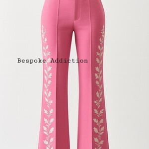 May include: Pink high-waisted pants with a flared leg. The pants feature a white leaf design on the sides and the text "Bespoke Addiction". The pants are displayed on a mannequin.