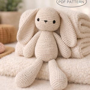 Può includere: Una bambola a forma di coniglio all'uncinetto di colore beige chiaro. Il coniglio ha lunghe orecchie, un piccolo naso e occhi neri ed è seduto su una morbida coperta strutturata. Il testo "PDF PATTERN" è in un ovale sopra il coniglio.
