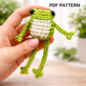 May include: A crocheted frog toy in green and white, with black eyes. The frog is held in a hand, and the text "PDF PATTERN" is visible. The frog has long legs and arms. A handmade item.