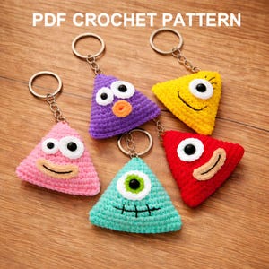 May include: A collection of handmade crochet triangle keychains in various colors, including pink, purple, yellow, red, and teal. Each keychain features a unique face design with googly eyes and smiling or frowning mouths. The text "PDF CROCHET PATTERN" is at the top.