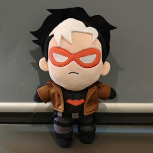 Peut inclure: Une poupée en peluche représentant un personnage aux cheveux noirs avec une mèche blanche, portant un masque rouge, une veste marron sur un t-shirt noir avec un emblème de chauve-souris rouge, et un pantalon sombre avec des sangles noires.