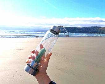 Puede incluir: Una botella de agua de vidrio transparente con una tapa plateada y un cristal verde en su interior. La botella tiene una correa negra unida a la tapa. La botella se está sosteniendo en el aire contra un fondo de playa y océano.