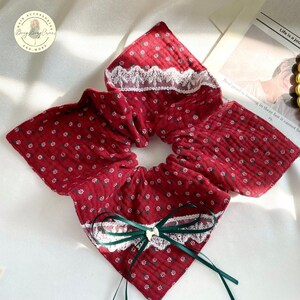 May include: A burgundy scrunchie with a floral pattern and white lace trim. The scrunchie has a matching fabric bow with green ribbon and a small white bow. The scrunchie is made of a soft, textured fabric.