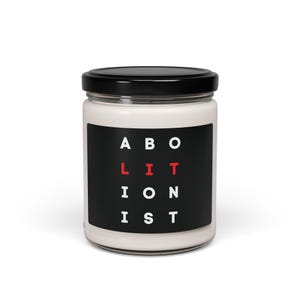May include: A white candle jar with a black lid and a black label that says "ABOLITION IS LIT" in white and red letters.