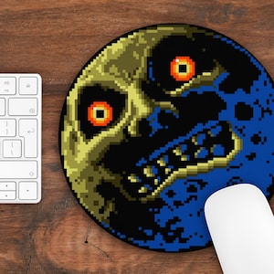 Op de afbeelding: Een ronde muismat met een gepixelde afbeelding van een gele en blauwe maan met een schedelgezicht en rode ogen. De maan is op een zwarte achtergrond.