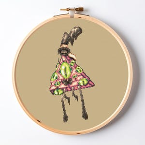 May include: A cross-stitch embroidery in a round wooden hoop featuring a pixelated creature with a pink, triangular body covered in multiple bright green eyes. The creature has a dark, curved top appendage and two thin, dark, dangling legs.