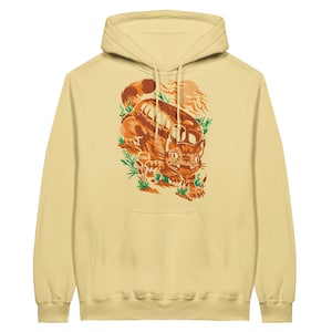 May include: A yellow hoodie with a cartoon illustration of a cat-like creature with large ears and a toothy grin. The creature is walking through a forest with a large, round object behind it. The illustration is in shades of orange, brown, and green.