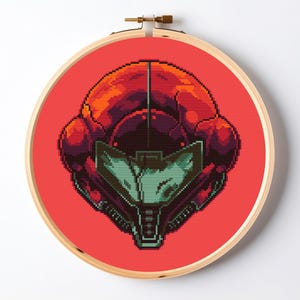 May include: A circular wooden embroidery hoop containing a cross-stitch piece featuring a pixelated, stylized helmet design. The helmet is rendered in shades of red, orange, and dark purple, with a central visor in light green and dark gray.