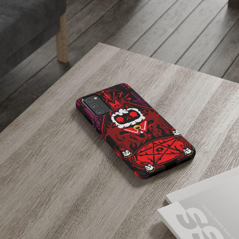 Op de afbeelding: Een zwarte telefoonhoes met een rode, zwarte en witte illustratie van een cartoonfiguur met een kroon en een staf. Het figuur is omringd door een pentagram en kaarsen. De hoes heeft een camera-uitsparing en een uitsparing voor de knoppen van de telefoon.