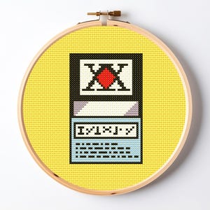 Puede incluir: Un bastidor de bordado circular de madera que contiene un diseño de punto de cruz sobre un fondo de tela amarillo brillante. El diseño presenta un borde rectangular negro con un cuadrado blanco que contiene un número romano X negro y un rombo rojo.