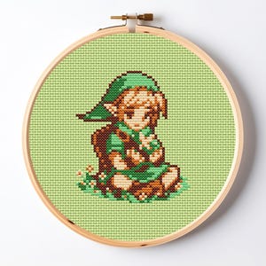 May include: A cross-stitch embroidery piece in a round wooden hoop featuring a pixelated character with blonde hair wearing a green tunic and cap, sitting on a patch of green grass with small white flowers against a light green fabric background.