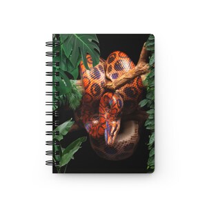 Puede incluir: Un cuaderno de espiral con una ilustración en la portada de una serpiente de patrones naranjas y negros enroscada en una rama de árbol marrón, rodeada de frondosas hojas tropicales verdes sobre un fondo negro sólido.