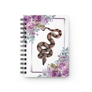 Könnte beinhalten: Ein spiralgebundenes Notizbuch mit weißem Einband, das in der Mitte eine Illustration einer braun-beigen Schlange zeigt. Das Motiv ist von einem dünnen violetten rechteckigen Rahmen umgeben und mit violetten Rosen und grünem Blattwerk verziert.