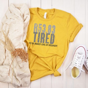 Può includere: Una maglietta gialla senape con la scritta "53.83 Tired to the highest level of specificity" stampata in grigio. La maglietta è abbinata a un cardigan di maglia bianco e scarpe da ginnastica bianche.