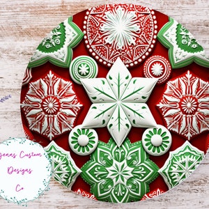 May include: A red, white, and green decorative plate with a snowflake and floral design. The plate features a central white star and various green and white floral shapes. The plate has the text "Jenns Custom Designs Co."