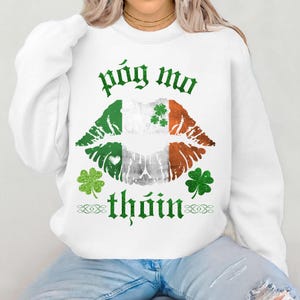 May include: White sweatshirt with a graphic design featuring a kiss with the Irish flag colors and shamrocks. The text "póg mo thóin" is printed in green above and below the lips.