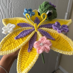 May include: A crocheted yellow lily flower with purple and green accents. The lily has a green crocheted frog sitting on top of it.