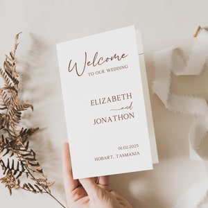 May include: A white wedding invitation card with the text "Welcome to our wedding" in a cursive font. The names "Elizabeth and Jonathon" are printed below, along with the date "01.02.2025" and the location "Hobart, Tasmania".