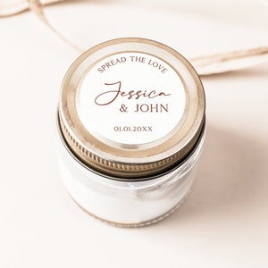 May include: A small glass jar with a silver lid. The lid has a white label with the text "SPREAD THE LOVE" above the names "Jessica & JOHN" and the date "01.01.20XX" in a brown script. The jar is filled with a white substance.