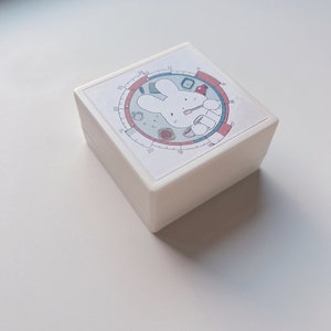 Könnte beinhalten: Weiße quadratische Box mit einem kreisförmigen Design, das ein Cartoon-Kaninchen zeigt, das eine Zahnbürste hält. Das Design enthält die Zahlen 1 bis 24 um den Kreis herum.