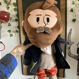 May include: A plush doll depicting a bearded man in a navy coat, orange trousers, and blue shoes. The doll has a brown beard and hair, with a neutral expression. The doll is a whimsical representation of a historical figure.