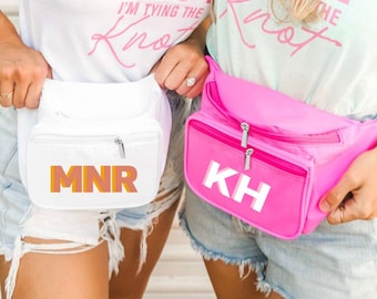 May include: Two white fanny packs with zippers and personalized initials. The fanny pack on the left has the initials "MNR" in orange and pink letters. The fanny pack on the right has the initials "KH" in white letters.