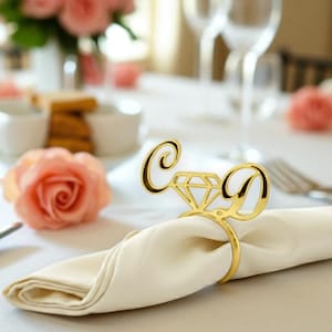 May include: A gold napkin ring featuring a diamond design and the letters "C" and "D". The ring holds a folded white napkin. A pink rose and other table settings are visible in the background.