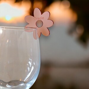 Può includere: Un segnabicchiere a forma di fiore rosa con il nome "Gracie" inciso su di esso. Il segnabicchiere è attaccato a un bicchiere di vino trasparente.