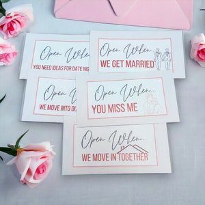 May include: Five white cards with pink and red text that say "Open When..." followed by different phrases. The phrases are "You need ideas for date night", "We get married", "We move into our place", "You miss me", and "We move in together".