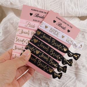 Puede incluir: Conjunto de cintas para el pelo negras y rosas con letras doradas. Las cintas para el pelo dicen "Bride" y "Team Bride". Las cintas para el pelo están empaquetadas en una tarjeta rosa con el texto "Siempre mira el lado de la novia de la vida".