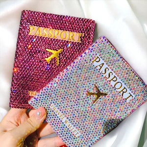 May include: Two passport covers with a sparkly, iridescent design. The covers are decorated with rhinestones and feature the word "PASSPORT" and a gold airplane graphic. The text "TRAVEL WALLET" is printed on the bottom of one cover.